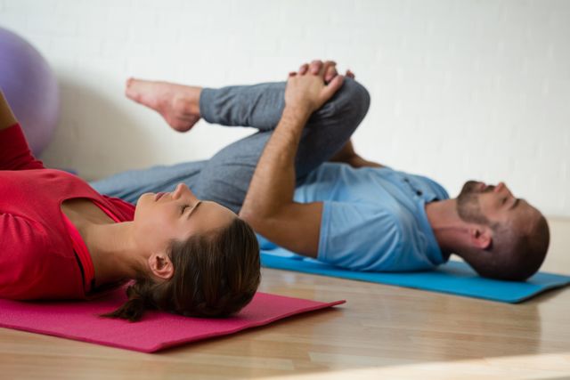 Instructor and student practicing yoga on mats in a studio. Both individuals are lying on their backs, engaging in a stretching exercise. Ideal for use in articles or advertisements related to fitness, wellness, yoga classes, and healthy lifestyles.