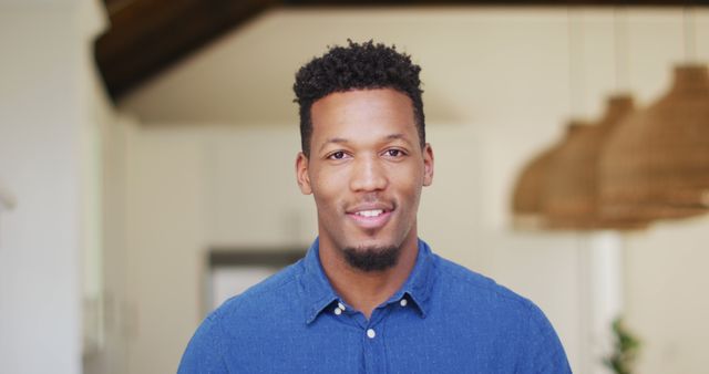 A confident African American man smiling while wearing a casual blue shirt indoors. The modern home setting with warm lighting creates a relaxed and comfortable atmosphere. This image is ideal for promotional materials, lifestyle blogs, social media content, and websites focusing on personal development or home living.