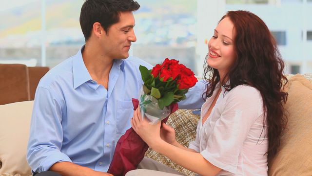 In an inviting living room, a man gives a bunch of red roses to his smiling wife, exuding feeling of love and affection. Perfect for ads focusing on romance, Valentine's Day, relationship advice, or social media posts celebrating partner appreciation and domestic bliss.