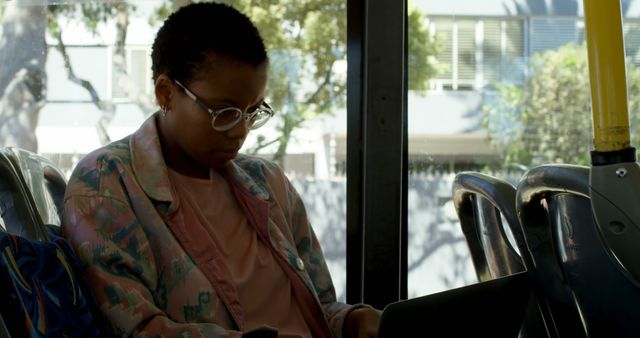 Young woman with short hair and glasses wearing a casual jacket, focused on her laptop while commuting on a bus. Window reveals cityscape outside suggesting daytime. Ideal for themes related to remote work, freelancing, productivity, urban living, public transportation, or digital lifestyle.
