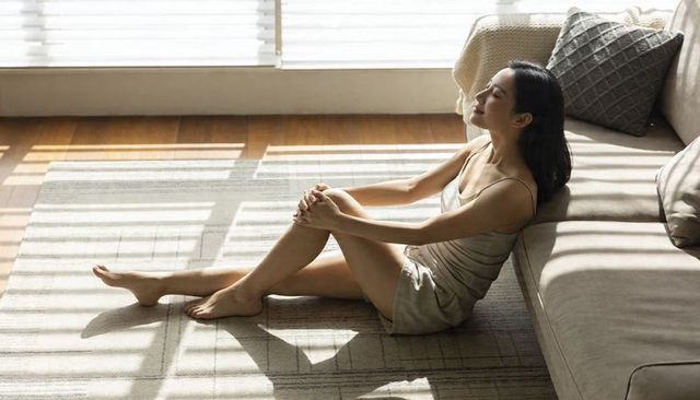 Asian woman relaxing in sunlit minimalist living room wearing satin camisole and shorts