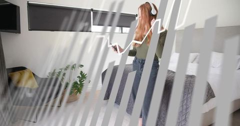 Redhead woman wearing white headphones holding smartphone enjoying music in minimalist bedroom