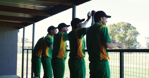 Cricket players leaning on dugout rail watching match from shaded dugout overlooking pitch