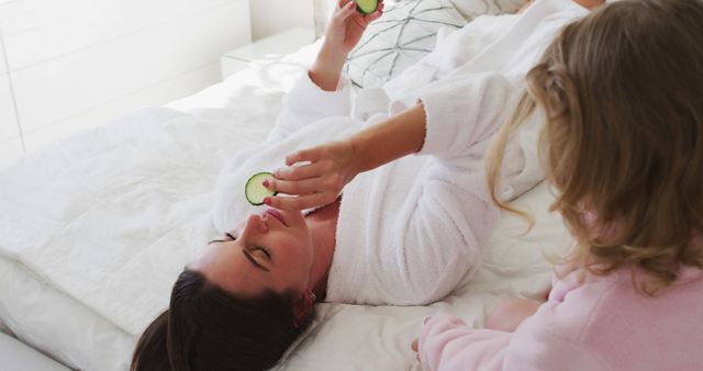 Woman lying on bed during a home spa day wearing a white bathrobe and holding cucumber slices on eyes. Another person beside in a pink bathrobe. Ideal for illustrating self-care, home spa day, beauty routine, relaxation, and wellness.