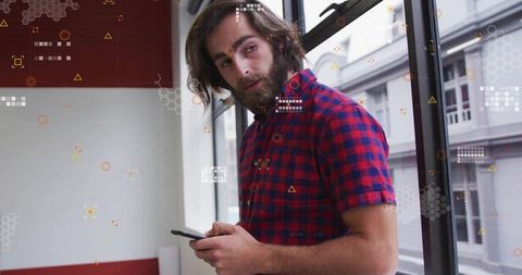 Bearded man holding smartphone glancing through urban window with geometric data overlay