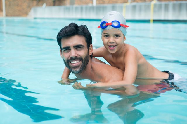 Father and son enjoying time together in a swimming pool. The child is wearing a swim cap and goggles, riding on the father's back. Both are smiling and having fun. Ideal for use in advertisements for family activities, leisure centers, summer vacations, and swimming lessons.