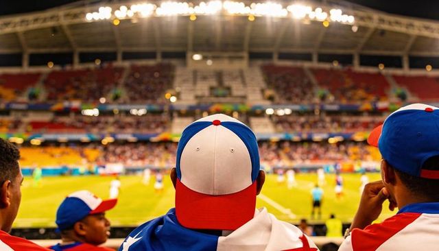 Fans watching night soccer match from stadium rail wearing red white blue caps and flags