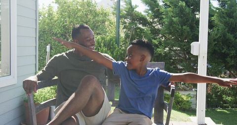 African american father and son playing airplane on sunny porch bench, joyful family moment