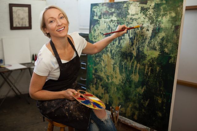 This image shows a woman happily painting on a canvas in an art studio. She is holding a paintbrush and a palette, wearing a casual outfit with an apron. This image can be used for promoting art classes, creative workshops, or hobby-related content. It is also suitable for illustrating articles about artistic expression, creativity, and personal fulfillment through art.