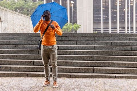 African american commuter holding blue umbrella calling on phone on rainy city steps