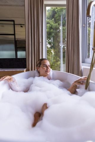 Woman relaxing in deep bubble bath by window in modern minimal bathroom for self-care