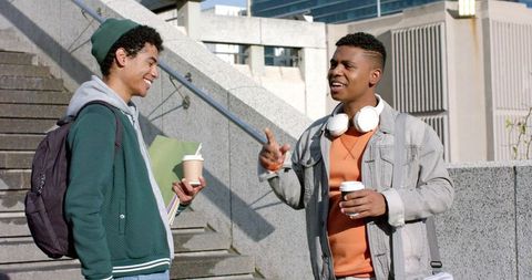 Diverse young men chatting on campus stairs drinking coffee and carrying backpacks