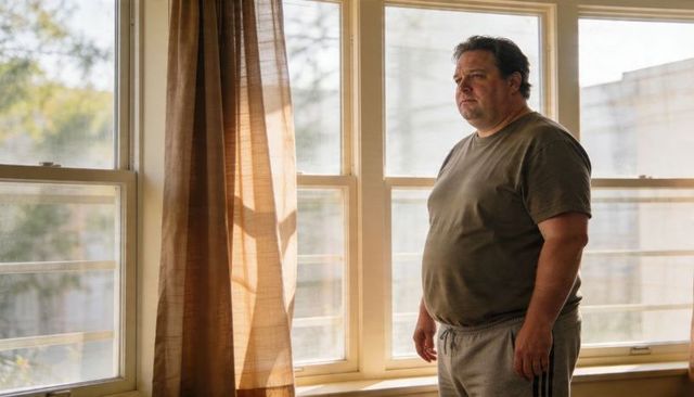 Midlife man gazing through sunlit window in olive tee and gray sweats, contemplative