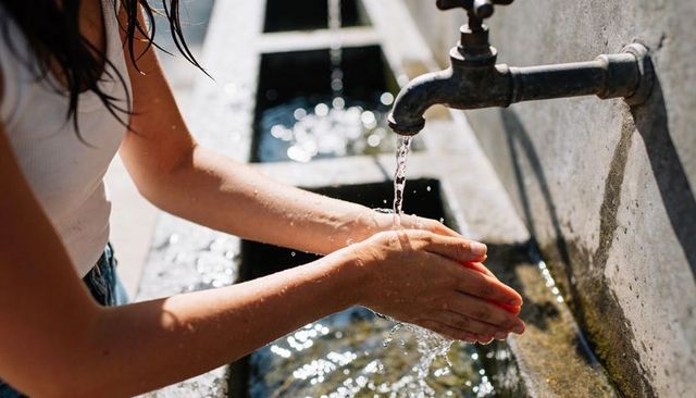 Washing hands under outdoor metal faucet with sunlight glinting on water droplets