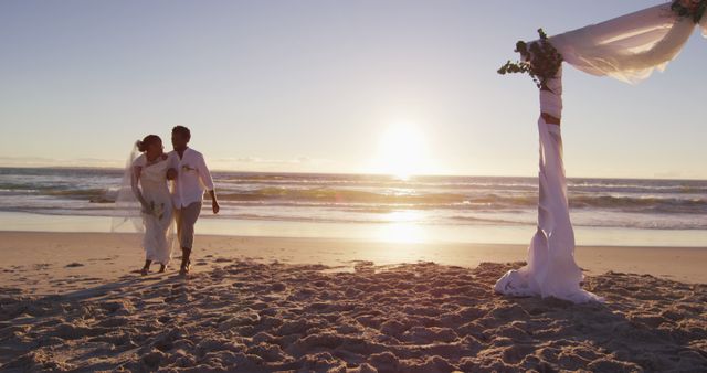 Bride and groom walking hand in hand on sandy beach with the sun setting and ocean waves in the background. Ideal for use in wedding themes, romance, travel promotions, or beach vacation advertisements.