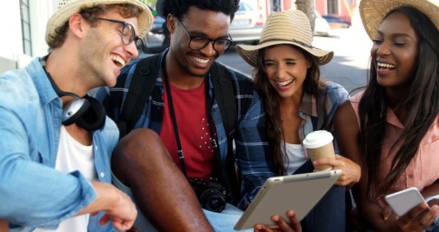 A diverse group of young adults is sharing a laugh while looking at a tablet, with copy space. Their casual attire and digital engagement suggest a relaxed social gathering in an urban setting.