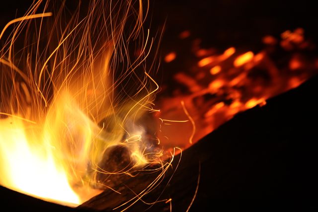 Closeup of campfire showcasing vibrant sparks and flames in dark outdoor setting, emitting warmth and light. Useful for themes of camping, nature, wilderness exploration, survival techniques, or illustrating warmth and coziness around fires.