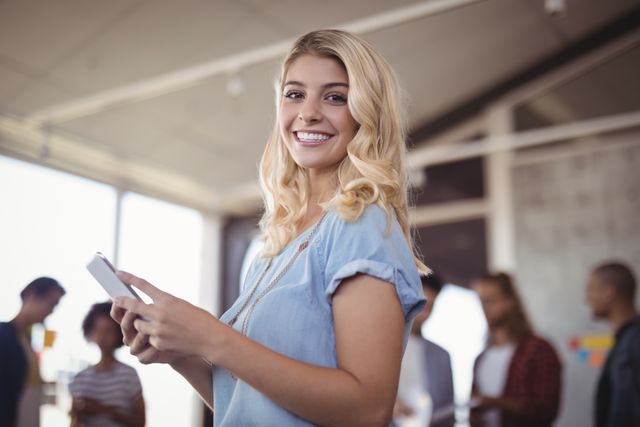 Young businesswoman smiling while holding a mobile phone in a creative office environment. Ideal for use in business, technology, and teamwork-related content. Perfect for illustrating modern workplace dynamics, startup culture, and professional communication.