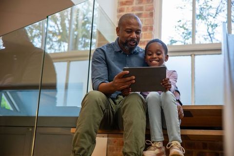 African american father sharing tablet with daughter on modern loft stairs, family bonding