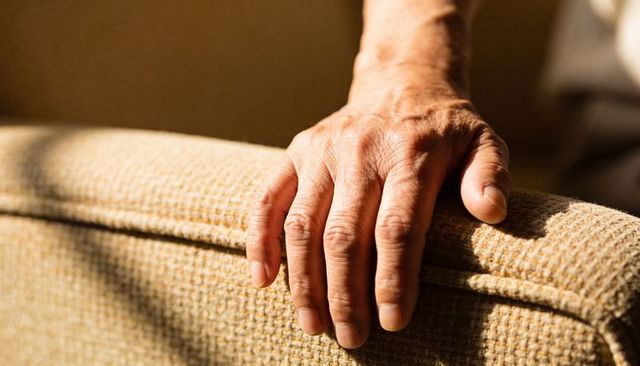 Resting senior male hand gripping beige sofa armrest, sunlight highlighting wrinkle detail