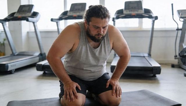 Overweight man kneeling panting after intense cardio workout in gym with treadmills