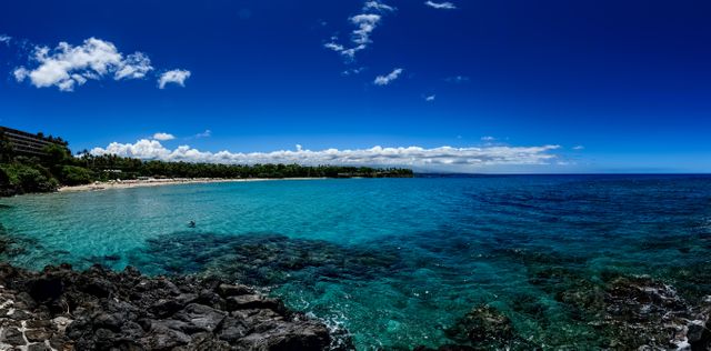 Azure waters meeting a rocky shore with lush greenery in the background and a clear blue sky overhead. Ideal for travel agencies, beach resort advertisements, vacation planning brochures, nature wallpapers, and summer-themed promotions.