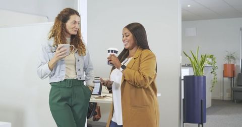 Two female colleagues chatting over coffee in bright modern open office lounge with plants