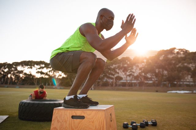 Fit african american man wearing face mask exercising outdoors jumping onto box. cross training for fitness at a sports field during coronavirus covid 19 pandemic.