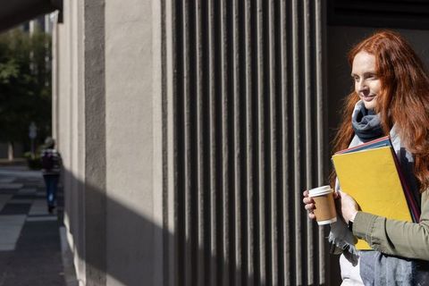 Sunlit red-haired student standing with coffee and colorful folders by fluted columns