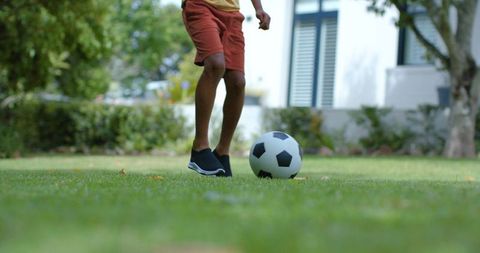 African american teen kicking soccer ball on sunny suburban lawn with garden and trees