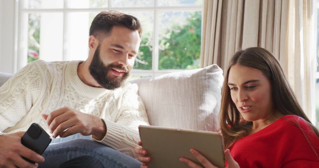 Image of happy caucasian couple siting on sofa and using tablet and smartphone. Domestic lifestyle and leisure at home.