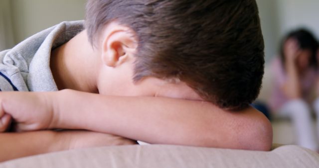 Sad boy resting his head on his arm, with distressed parent in the background. Suitable for articles and materials on family issues, mental health, psychological struggles in children, and conveying themes of domestic emotional challenges.