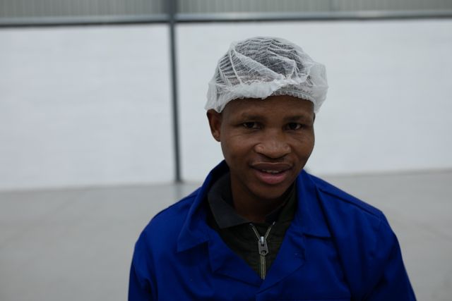 Portrait close up of an African American male worker in a factory workshop, wearing a hair net and blue overalls, looking to camera and smiling