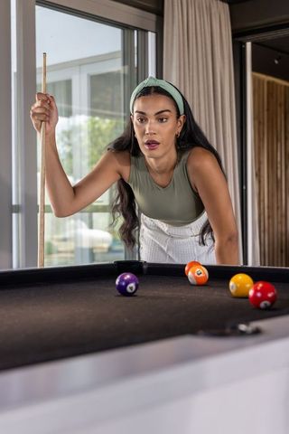 Young woman aiming cue on black pool table in modern sunlit lounge with patio view