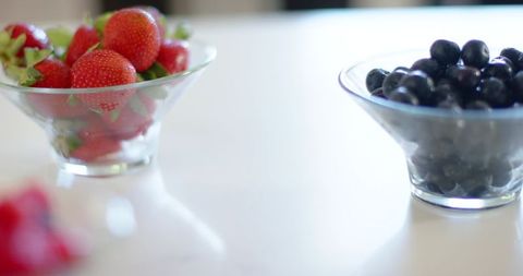 Fresh strawberries and blueberries in glass bowls on glossy white counter catching light
