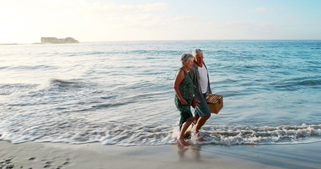 A senior couple is walking along a beach, holding a picnic basket and enjoying the gentle waves of the ocean under the sunset sky. Perfect for illustrating concepts of retirement, romance, togetherness, healthy lifestyle, peaceful holidays, and outdoor activities for elderly people.