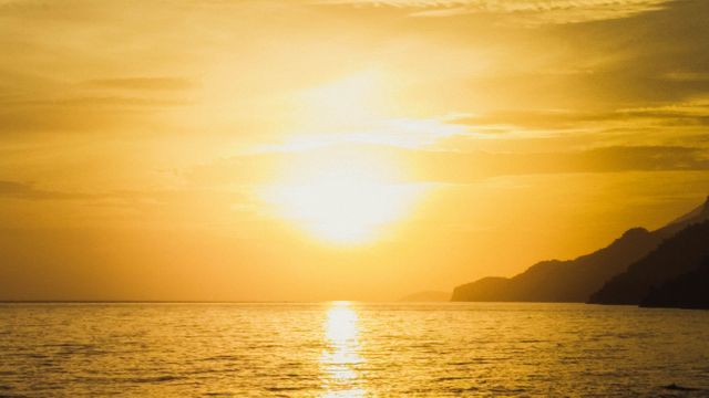 Image showcases tranquil golden sunset over calm ocean with distant hills. Ideal for travel promotions, relaxation themes, nature blogs, and background uses. Highlights peaceful scenery, making it suitable for printed materials, website headers and inspirational content.