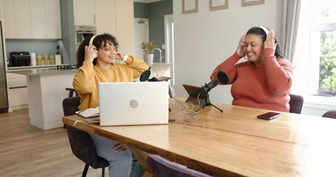 Diverse women recording podcast at home kitchen table wearing headphones and smiling