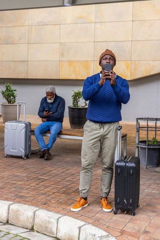 Urban traveler standing with black suitcase using smartphone outside transit hub, waiting