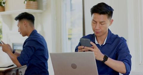Asian man balancing laptop work with smartphone use at home office