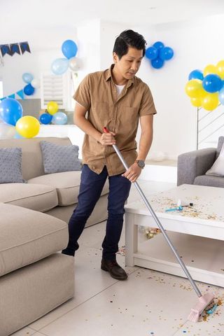Asian man sweeping confetti in living room after party with balloons and banner