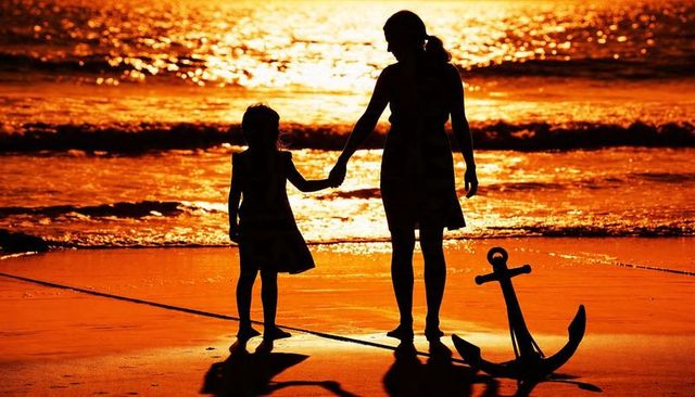 Mother and daughter holding hands on sunset beach with anchor silhouette