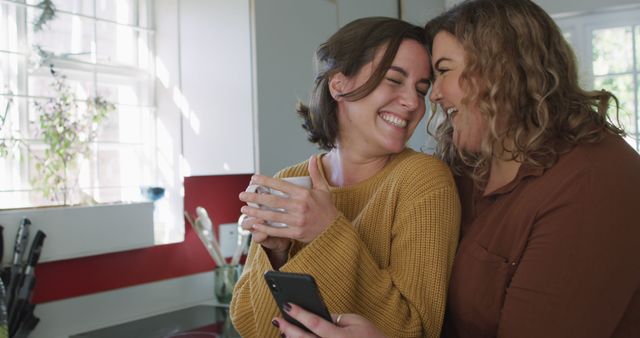 Happy caucasian lesbian couple embracing and using smartphone in kitchen. domestic life and leisure time.