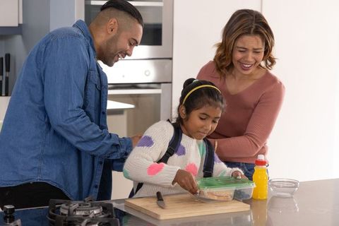Multicultural parents helping child prepare lunch while adjusting backpack on island