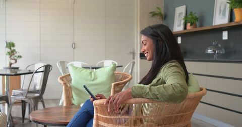 Indian woman relaxing in rattan chair using smartphone in cozy modern living room
