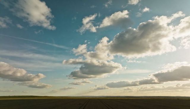 Sunlit cumulus clouds over plowed farmland with wide blue sky, contrails and green horizon