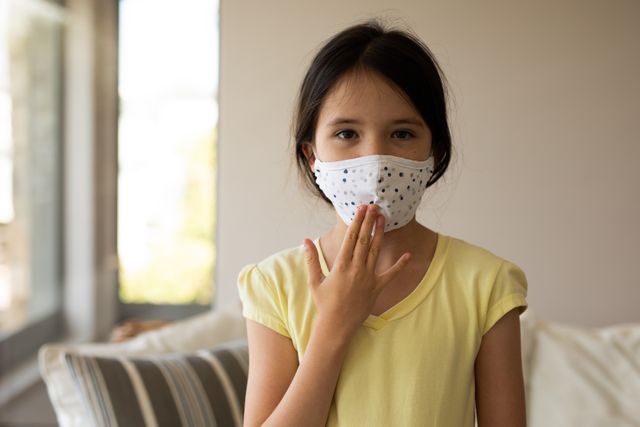 Little caucasian girl standing in front of the couch in their house while wearing a face mask. she is touching her facemask with her fingers.