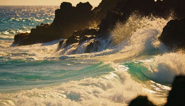 Crashing turquoise waves colliding with jagged rocks at golden hour