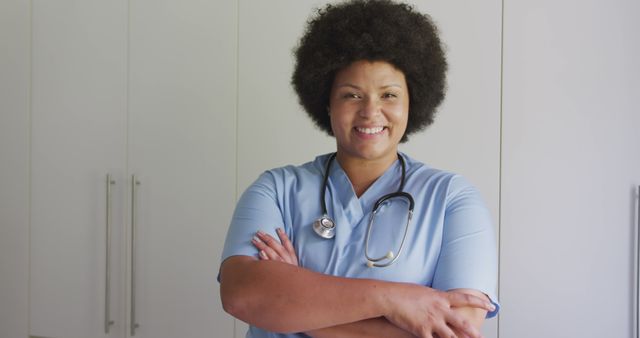African American nurse is standing, smiling and looking confident, standing in front of white cabinets. Wearing light blue scrubs and a stethoscope, she has her arms crossed. This image may be used on healthcare websites, medical advertisements, or in articles highlighting the healthcare profession.