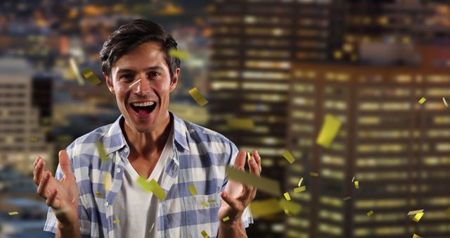 This image shows a young and excited man joyfully celebrating amidst gold confetti in a night-time urban backdrop featuring well-lit buildings. Great for illustrating themes of victory, celebration, success, or happiness in marketing, advertising, or social media posts about personal or professional milestones.
